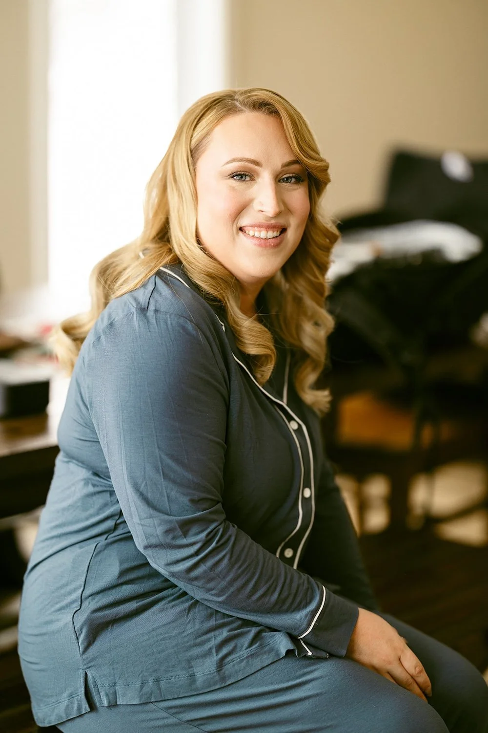 The bride smiles in her wedding day pajamas with make up and hair completed. 