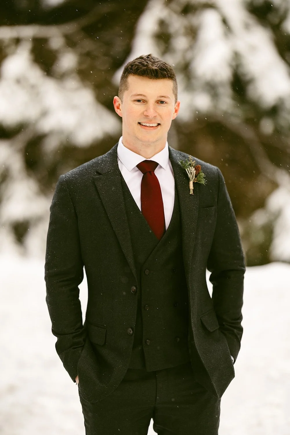 The groom stands in his black suit with the snowy evergreens and trail surrounding him. 