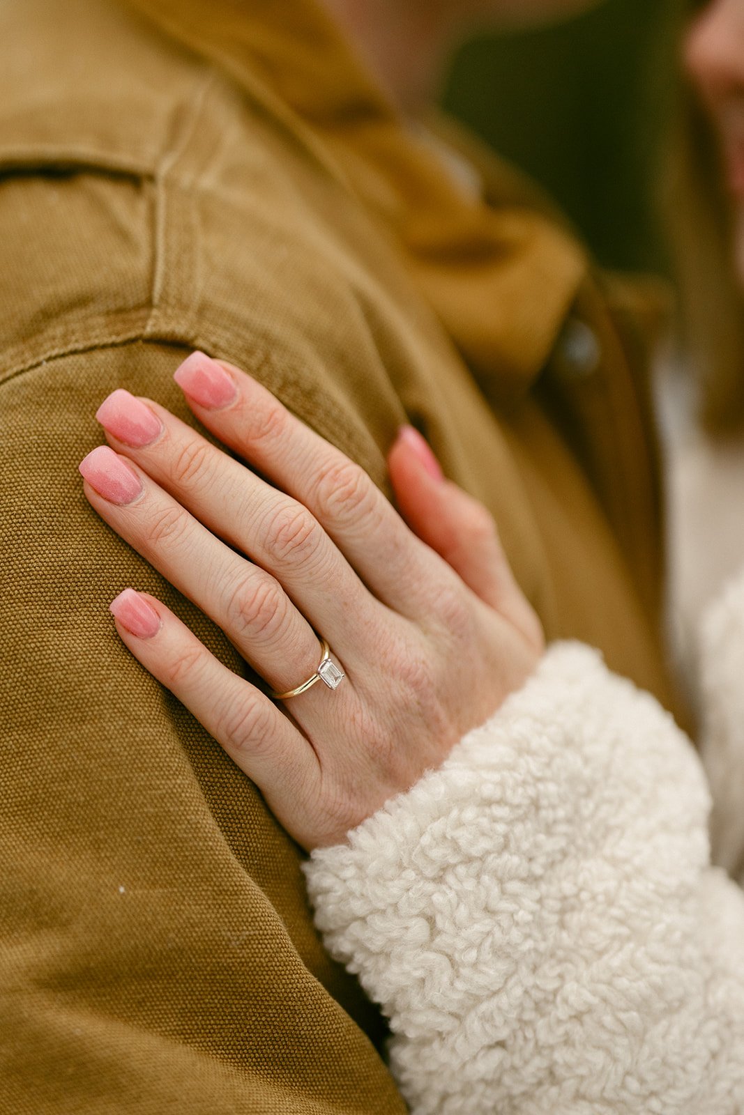 A close-up of a woman's hand with pink nails and an engagement ring resting on a person's shoulder wearing a brown jacket.