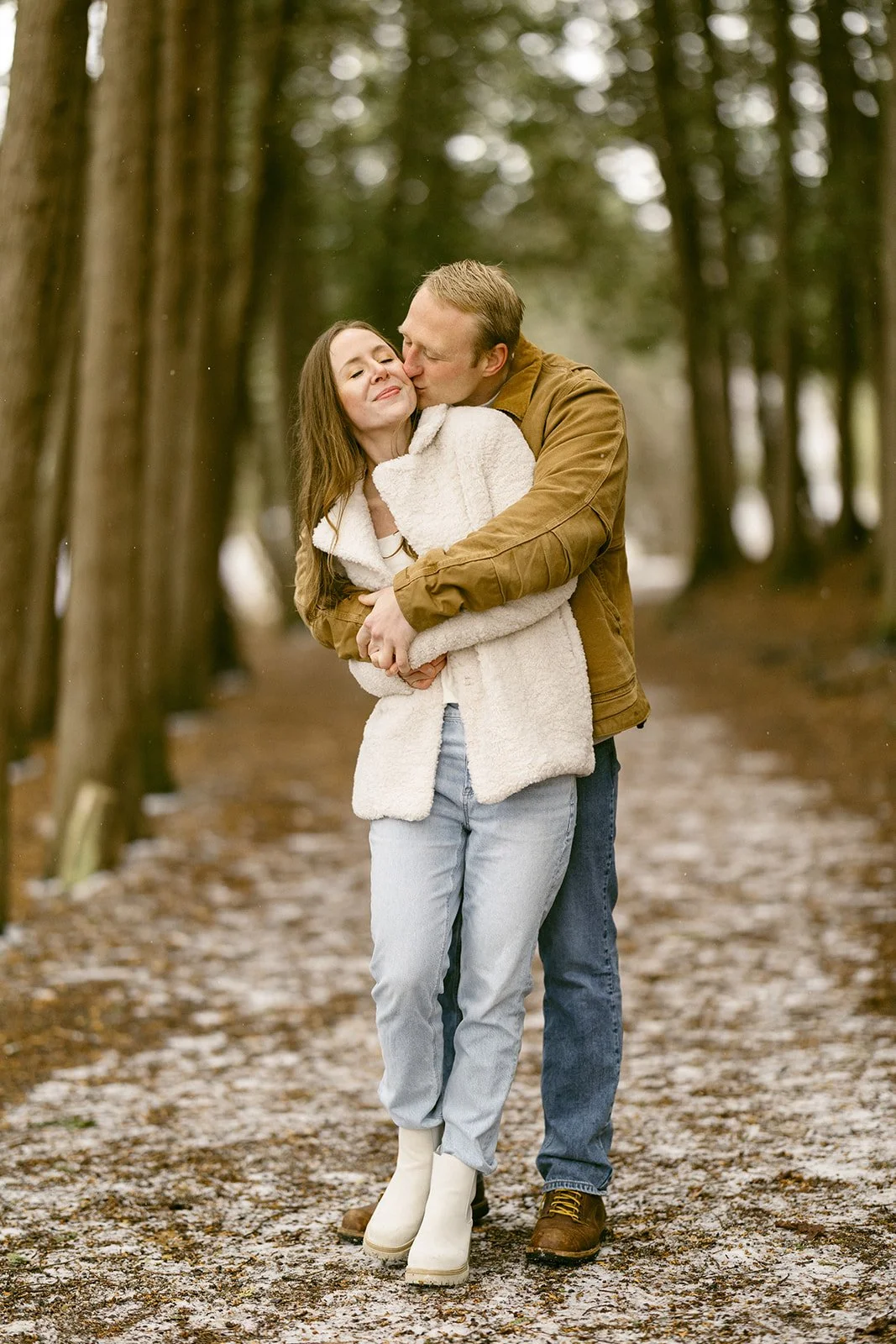 A couple stands on a leaf-covered path surrounded by tall, closely spaced trees in a forest for winter engagement photos at Green Lakes State Park