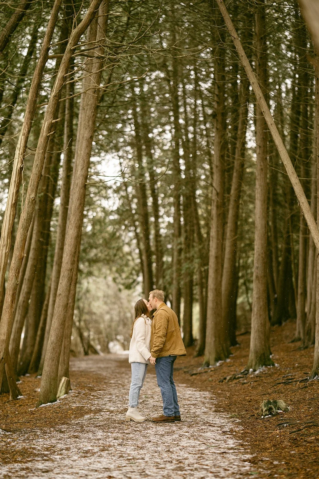 A couple stands on a leaf-covered path surrounded by tall, closely spaced trees in a forest for winter engagement photos at Green Lakes State Park