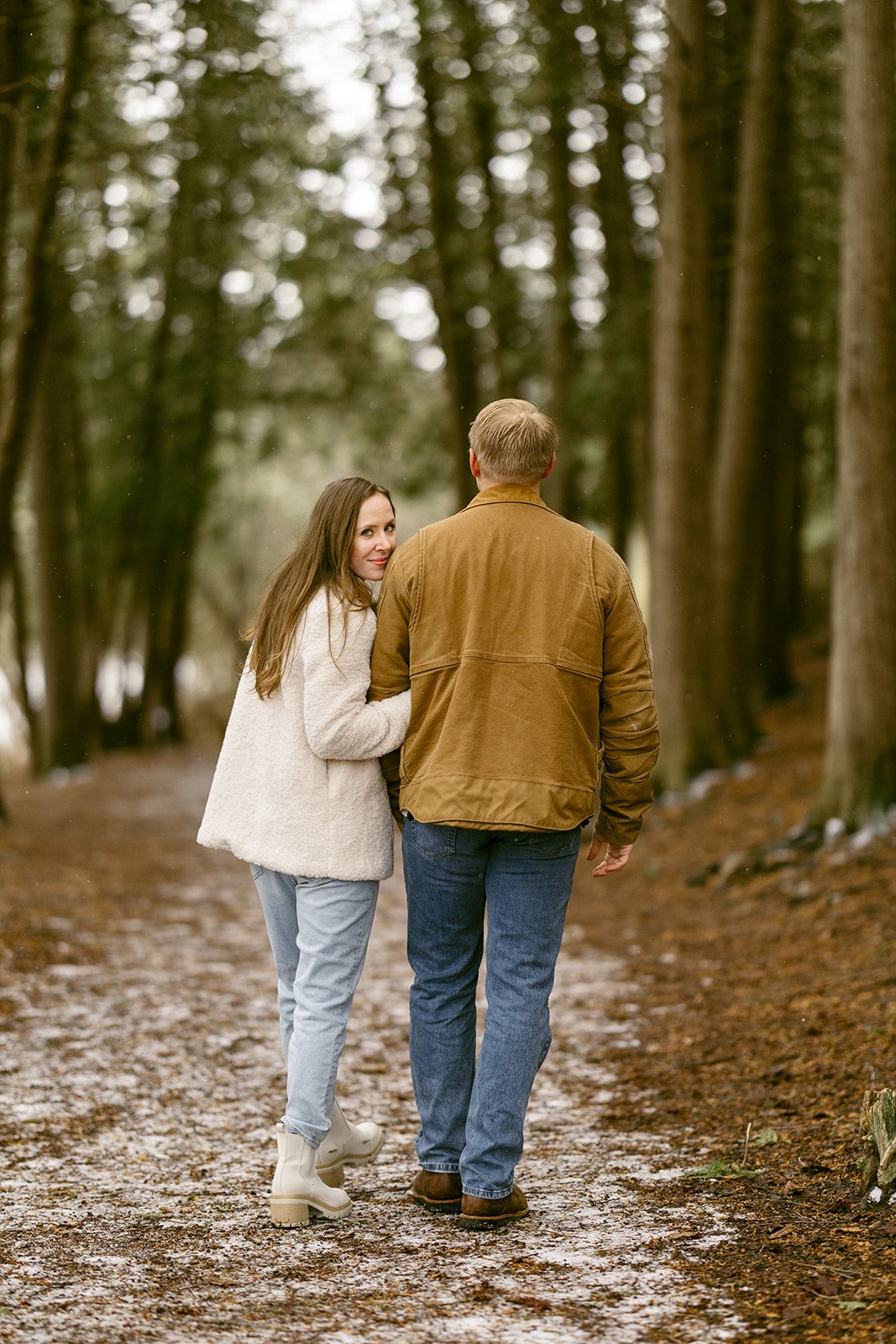 A couple stands on a leaf-covered path surrounded by tall, closely spaced trees in a forest for winter engagement photos at Green Lakes State Park