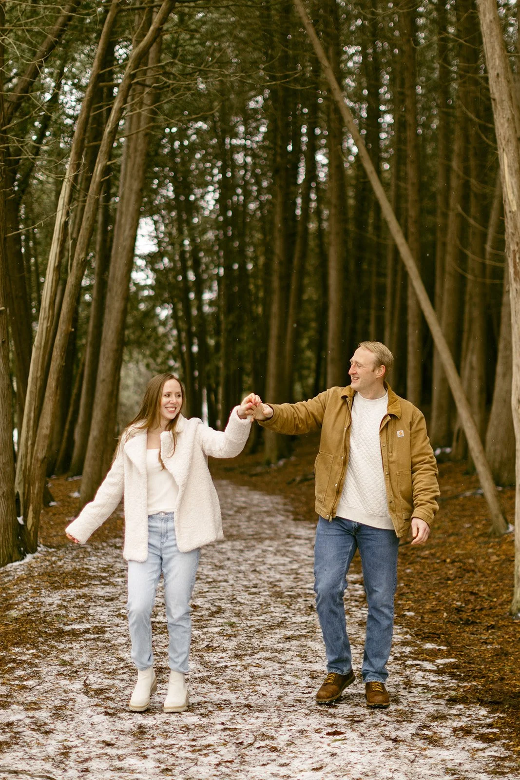 A couple stands on a leaf-covered path surrounded by tall, closely spaced trees in a forest for winter engagement photos at Green Lakes State Park