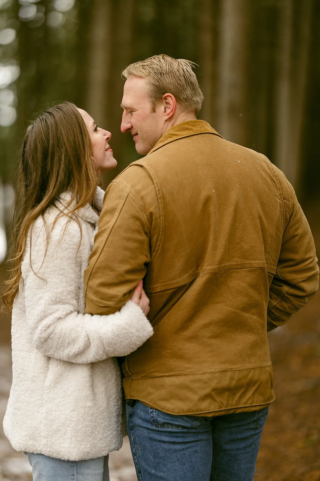 A couple stands on a leaf-covered path surrounded by tall, closely spaced trees in a forest for winter engagement photos at Green Lakes State Park