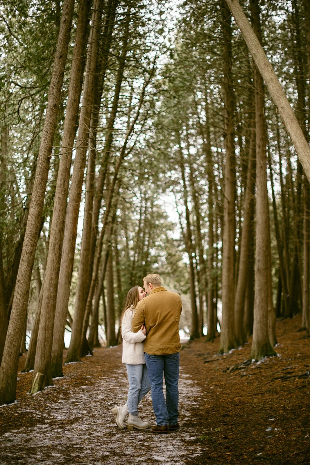 A couple stands on a leaf-covered path surrounded by tall, closely spaced trees in a forest for winter engagement photos at Green Lakes State Park