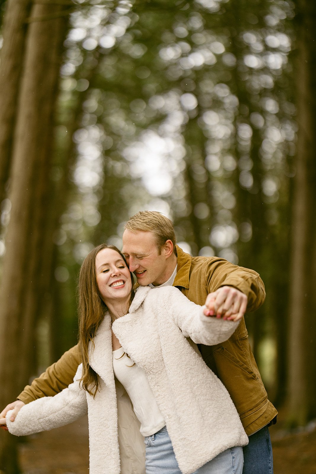 A couple stands on a leaf-covered path surrounded by tall, closely spaced trees in a forest for winter engagement photos at Green Lakes State Park