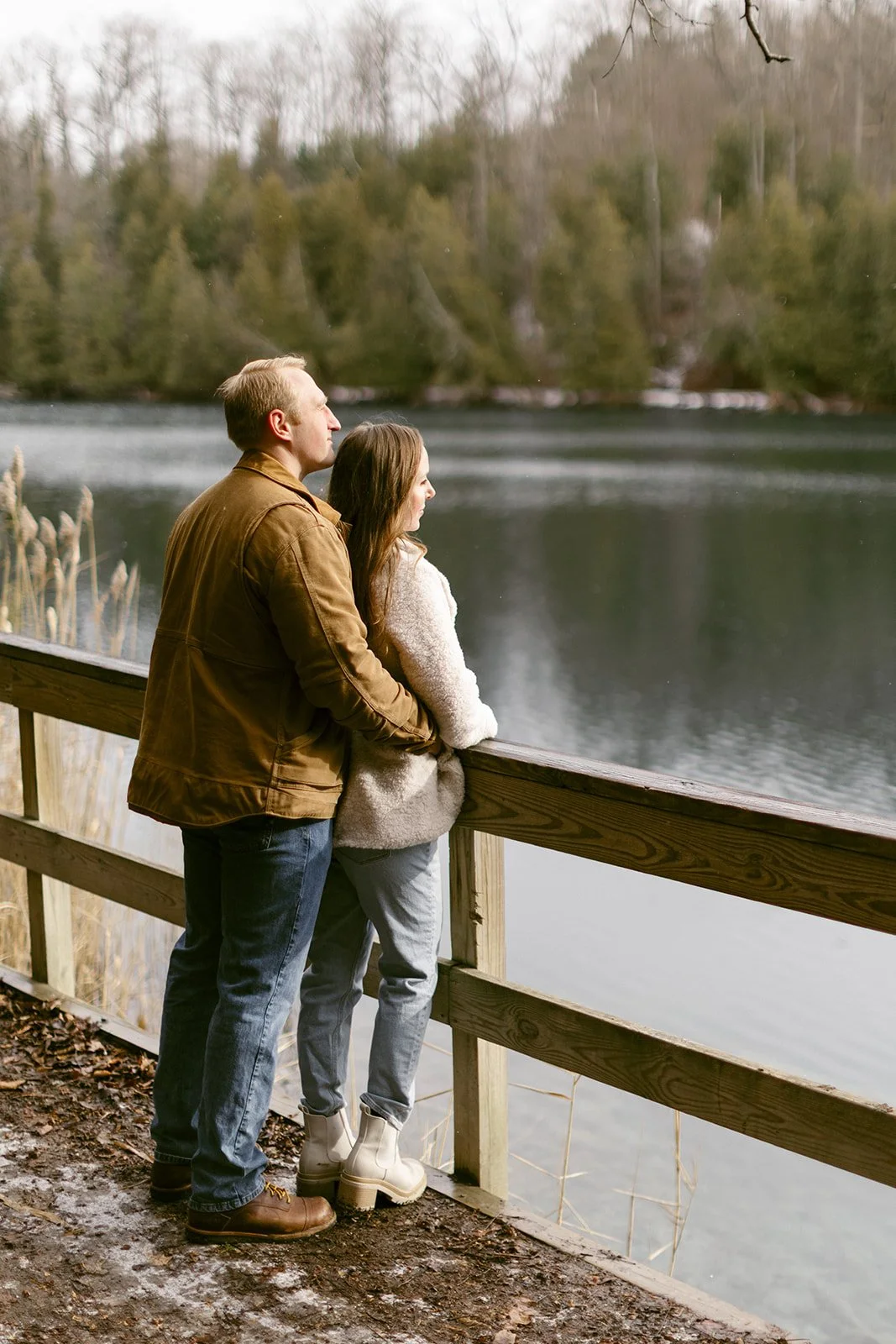 A couple stands side by side at a wooden railing, looking out over a calm lake surrounded by trees.