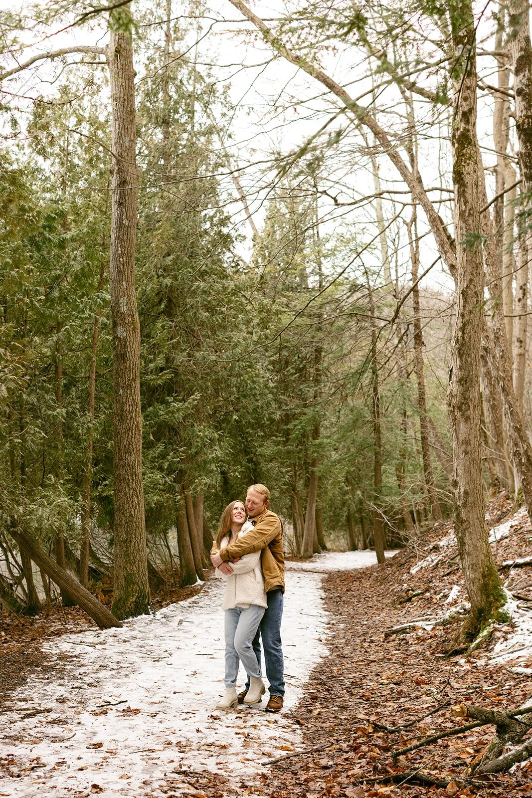 A couple stands close together on a snowy path in a forest, with the man gently leaning his forehead against the woman's temple.