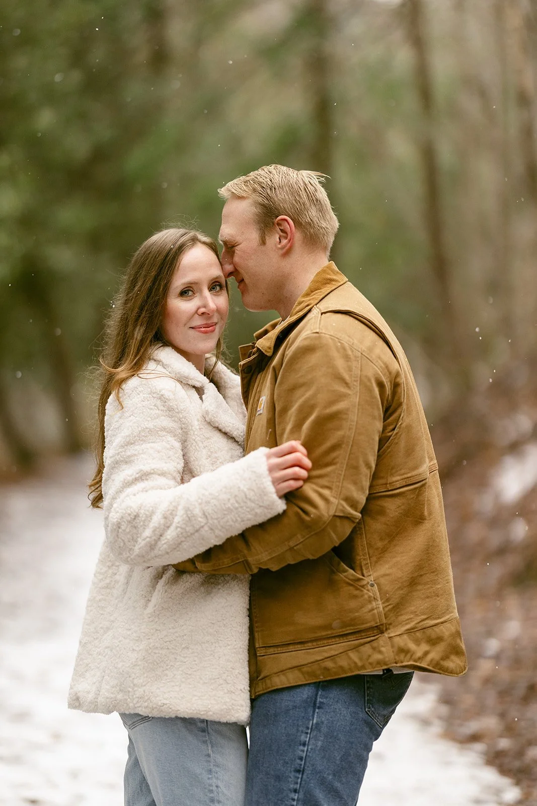 A couple stands close together on a snowy path in a forest, with the man gently leaning his forehead against the woman's temple.