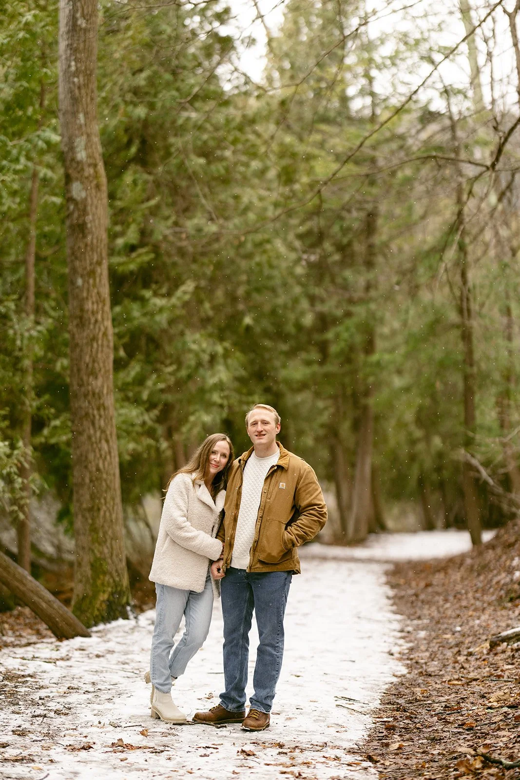 A couple stands side by side at a wooden railing, looking out over a calm lake surrounded by trees for winter engagement photos