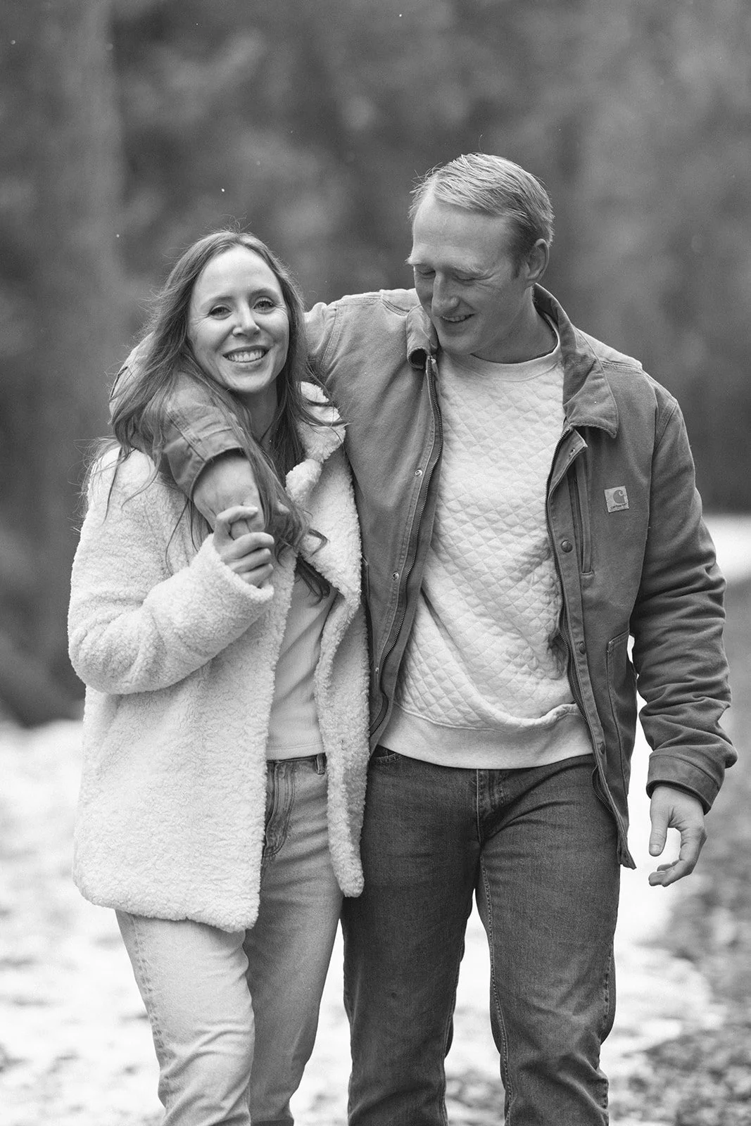 A couple stands side by side at a wooden railing, looking out over a calm lake surrounded by trees for winter engagement photos