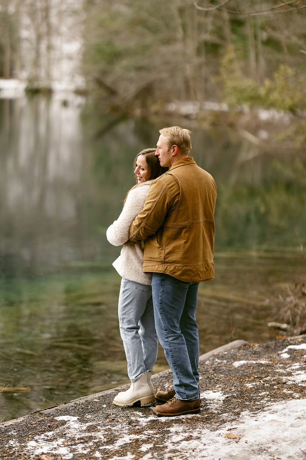 A couple stands side by side at a wooden railing, looking out over a calm lake surrounded by trees for winter engagement photos
