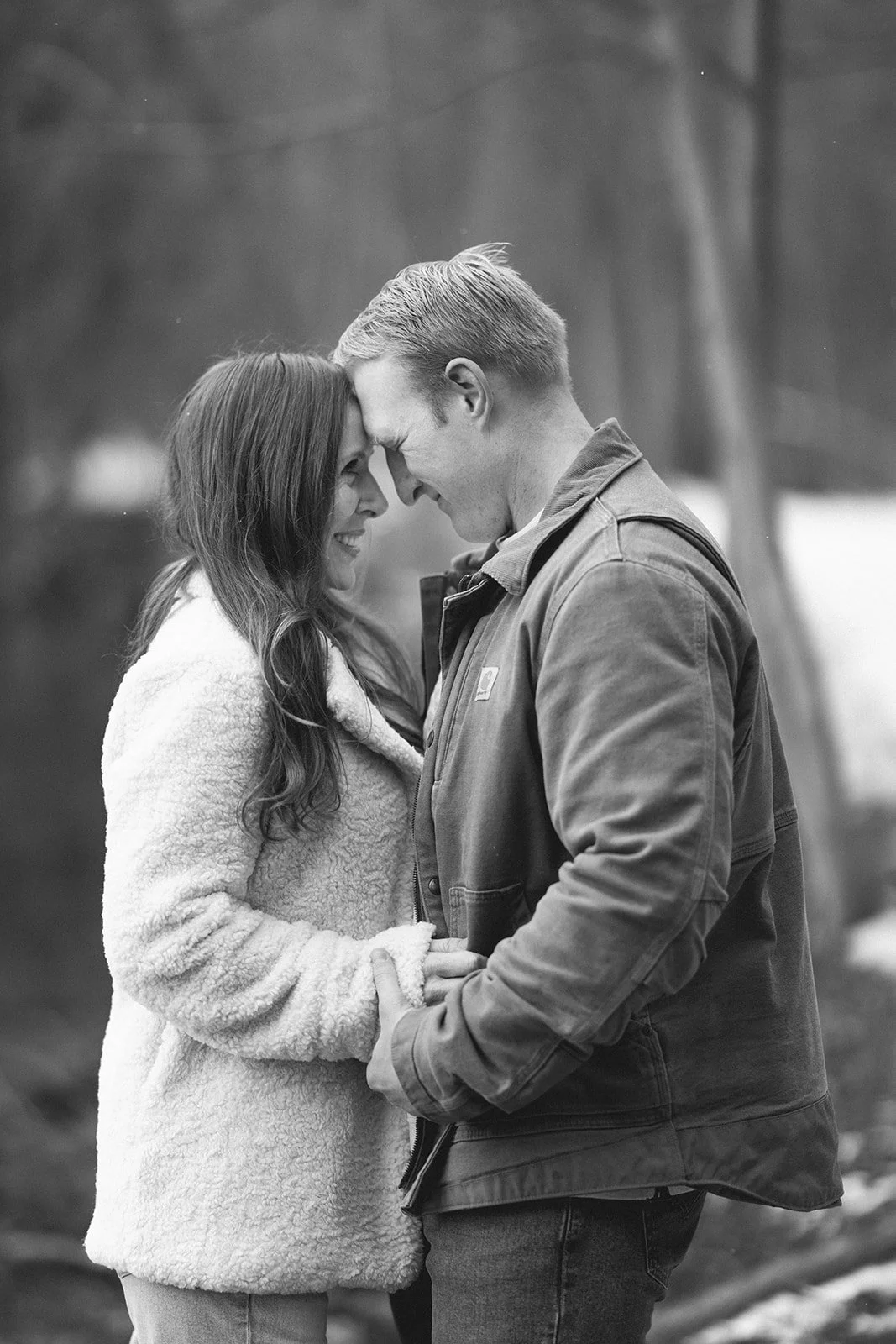 A couple stands side by side at a wooden railing, looking out over a calm lake surrounded by trees for winter engagement photos