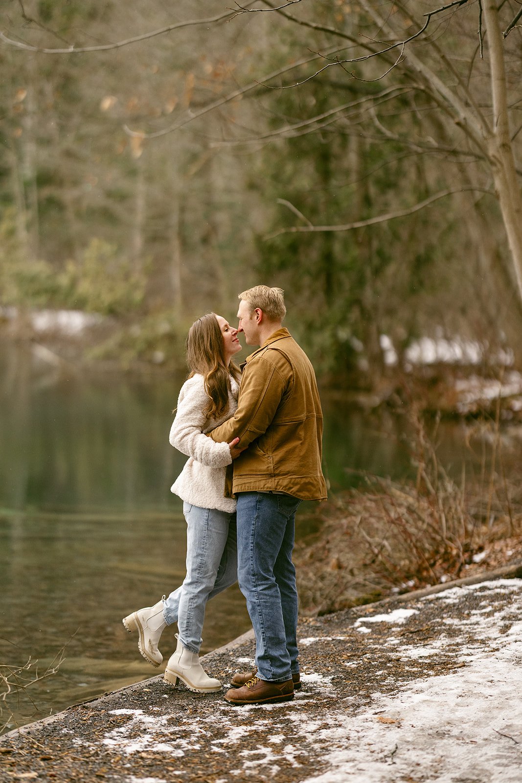 A couple stands side by side at a wooden railing, looking out over a calm lake surrounded by trees for winter engagement photos