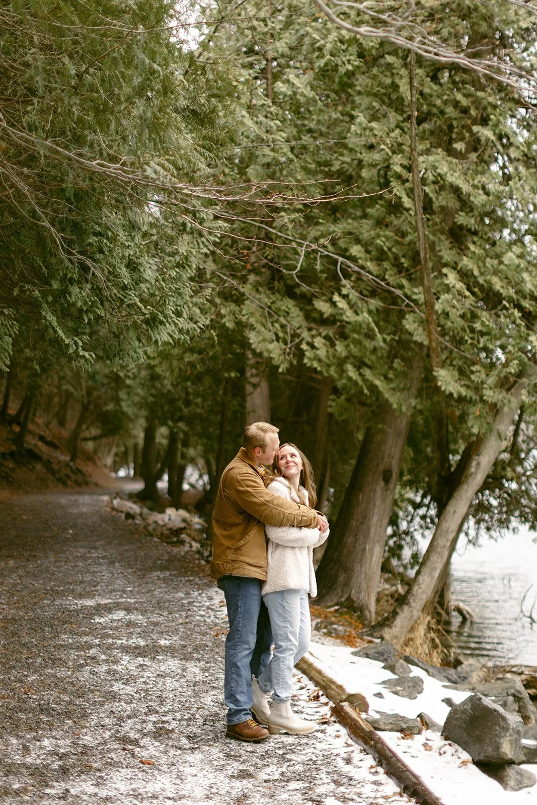 A woman in a white coat smiles while being hugged from behind by a man in a brown jacket.