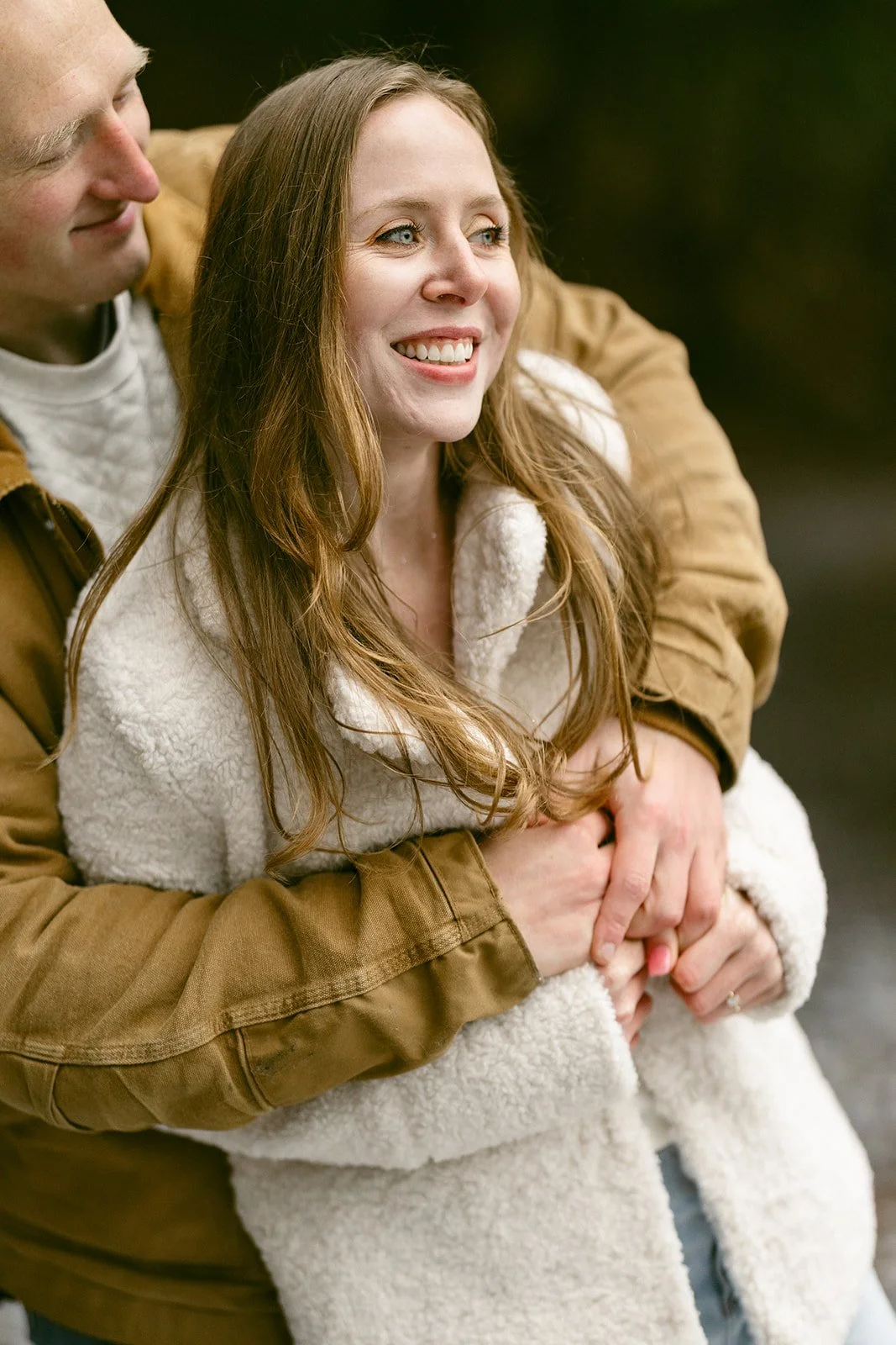 A woman in a white coat smiles while being hugged from behind by a man in a brown jacket.