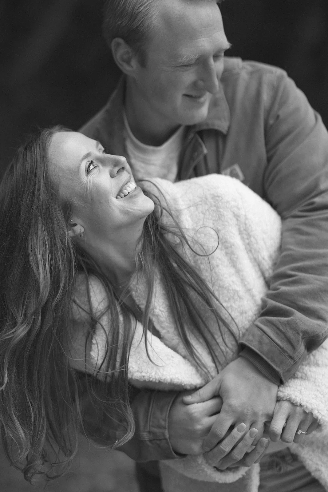 A woman in a white coat smiles while being hugged from behind by a man in a brown jacket.