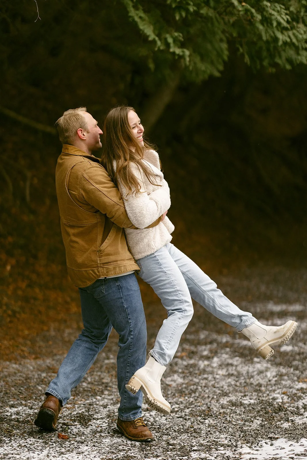 A couple stands embracing on a snowy path beside a lake, surrounded by tall trees.