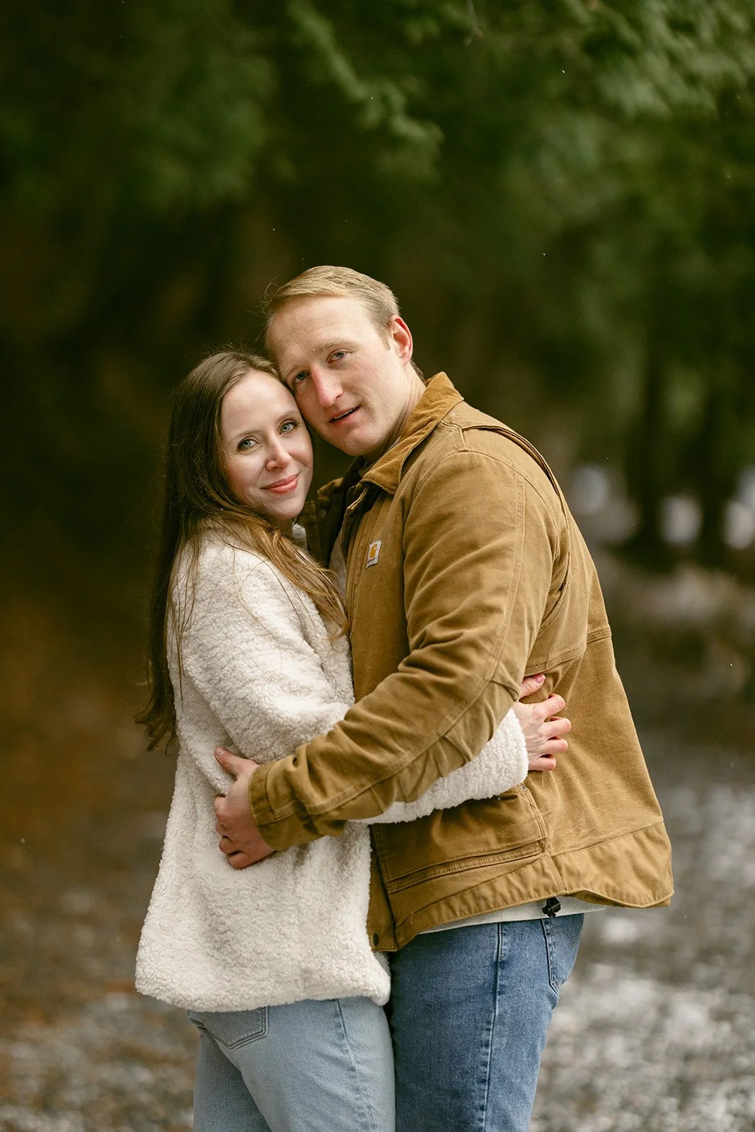 A couple stands embracing on a snowy path beside a lake, surrounded by tall trees.