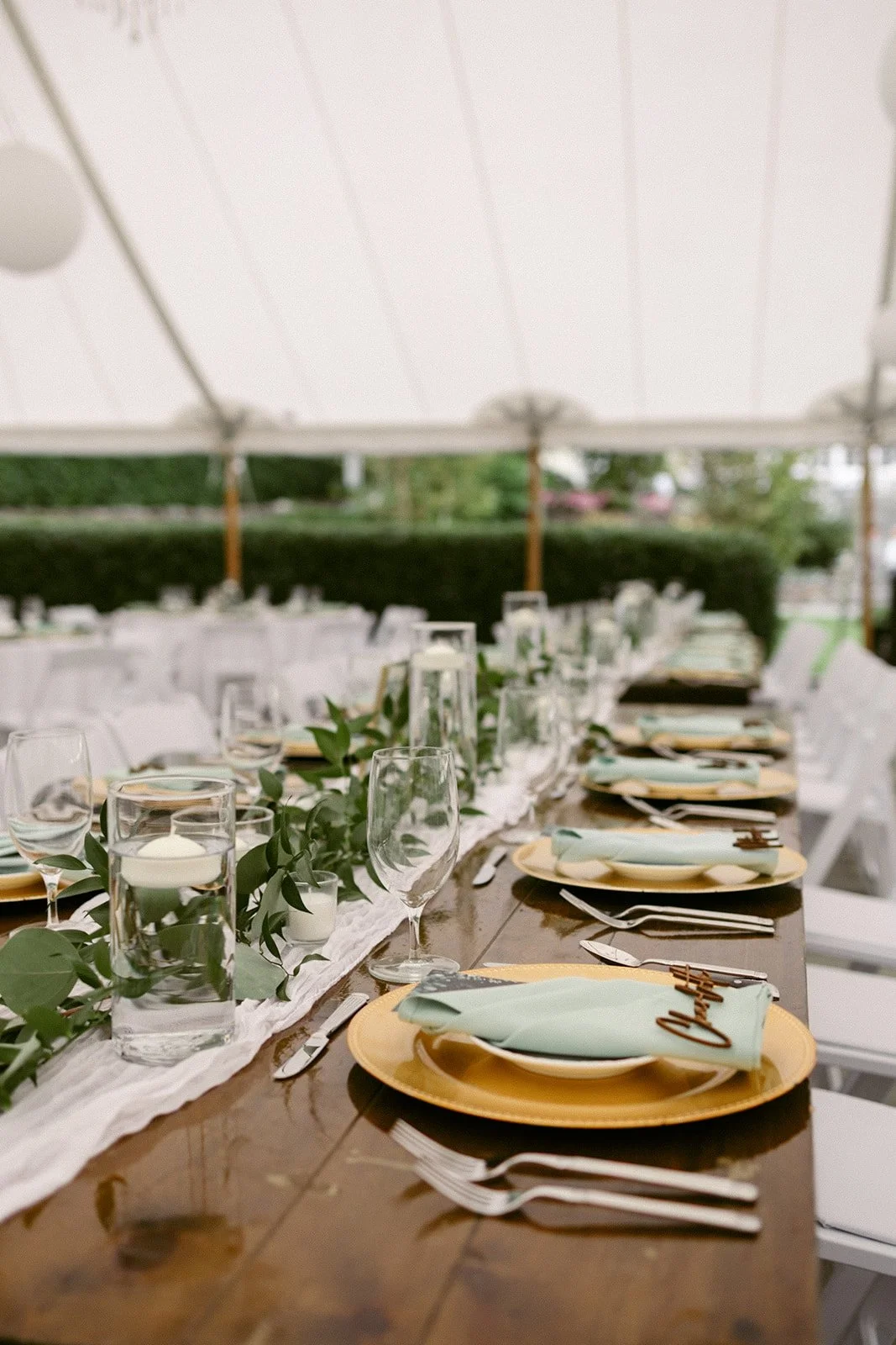 A long, wooden banquet table is set with plates, cutlery, glasses, and decorative greenery under a white tent for an outdoor event.