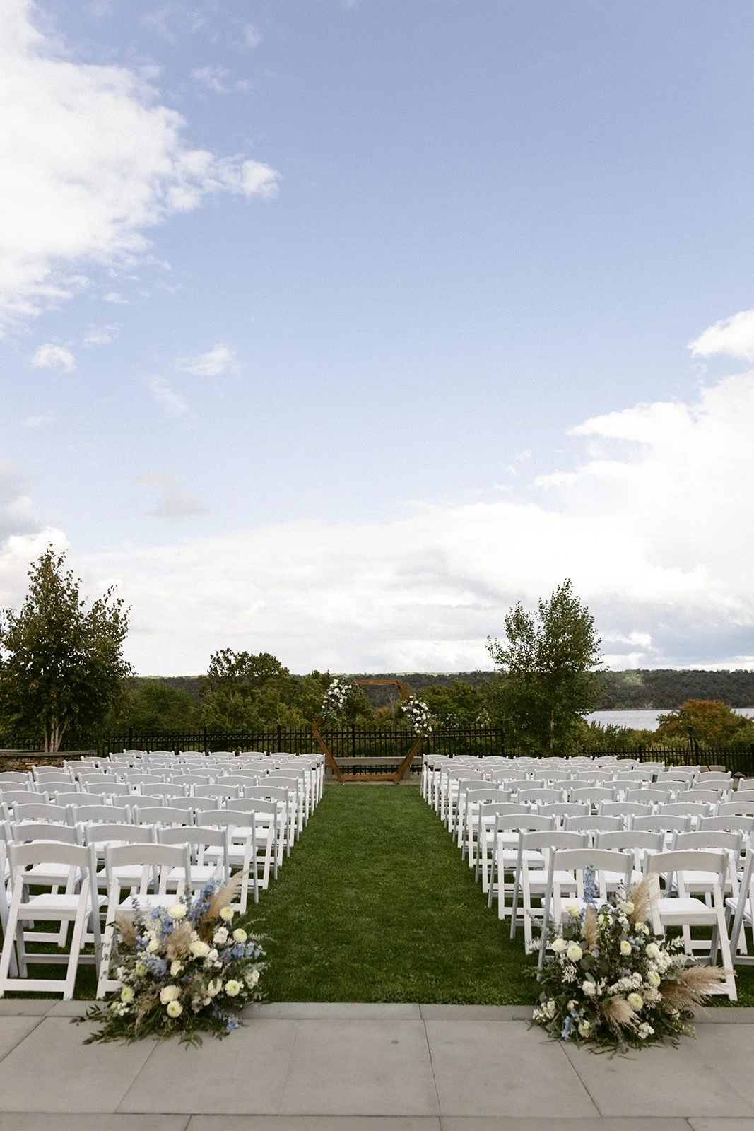 Rows of white folding chairs arranged outdoors on a lawn for a wedding ceremony, with floral arrangements at the aisle entrance and trees in the background at the Inn at Taughannock Falls