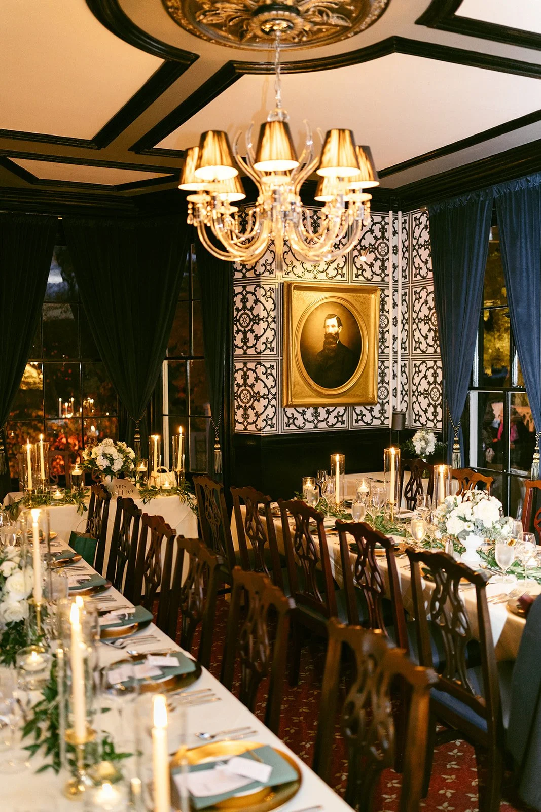 The Jones room at Inn at Taughannock Falls with long banquet tables, floral centerpieces, and candlelight, featuring a chandelier and a portrait on a patterned wall.