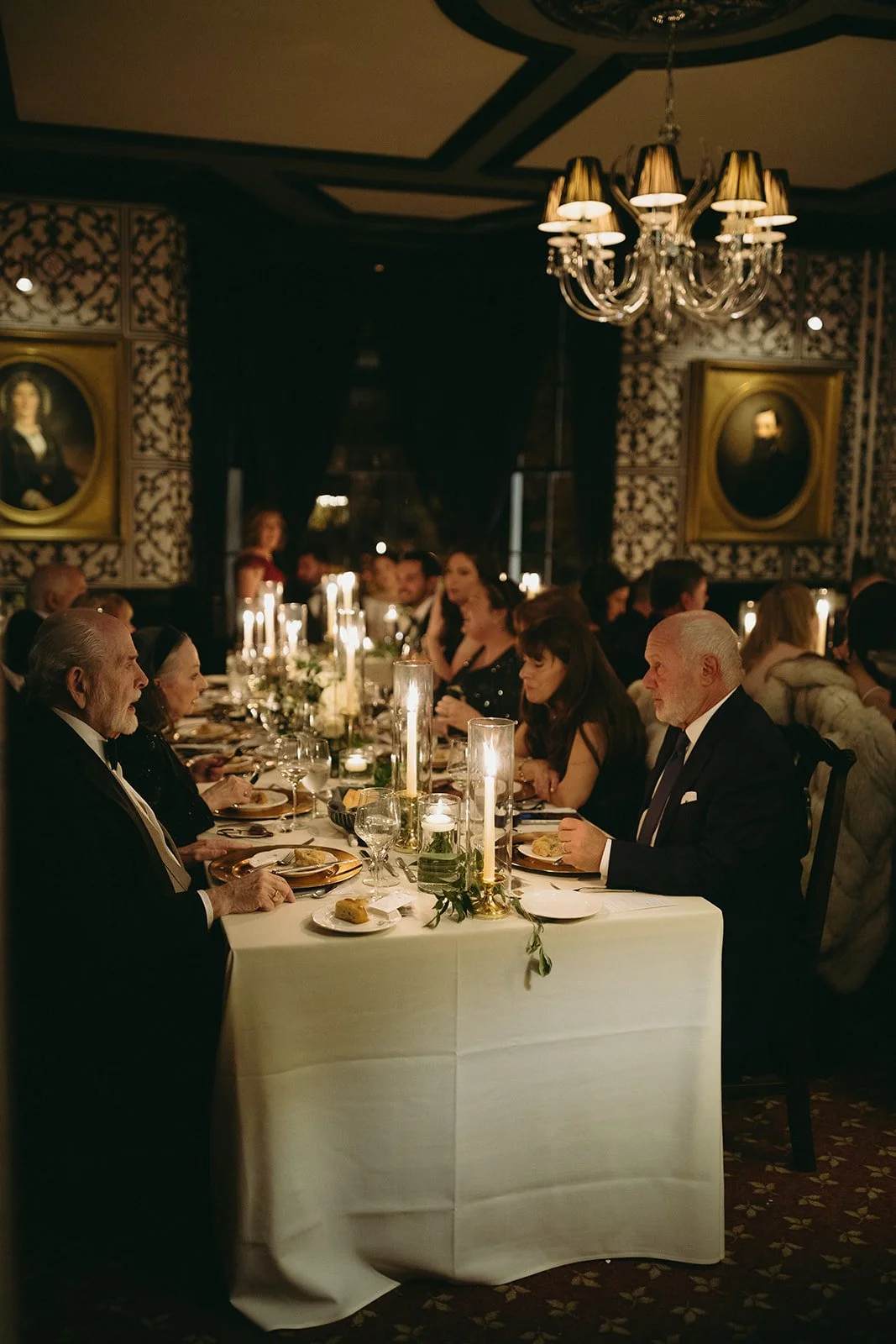 The Jones room at Inn at Taughannock Falls with long banquet tables, floral centerpieces, and candlelight, featuring a chandelier and a portrait on a patterned wall.