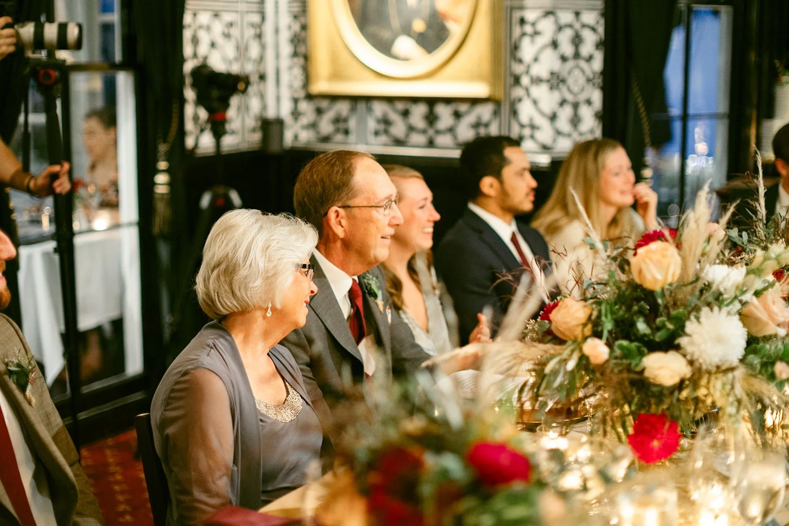 The Jones room at Inn at Taughannock Falls with long banquet tables, floral centerpieces, and candlelight, featuring a chandelier and a portrait on a patterned wall.