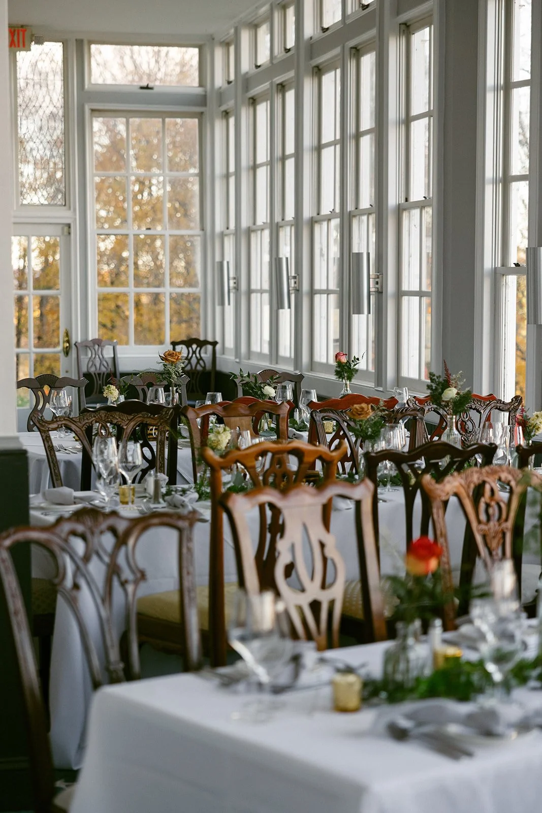 Elegant dining room with wooden chairs, white tablecloths, and tables set with glassware, cutlery, and floral centerpieces, beside large sunlit windows.