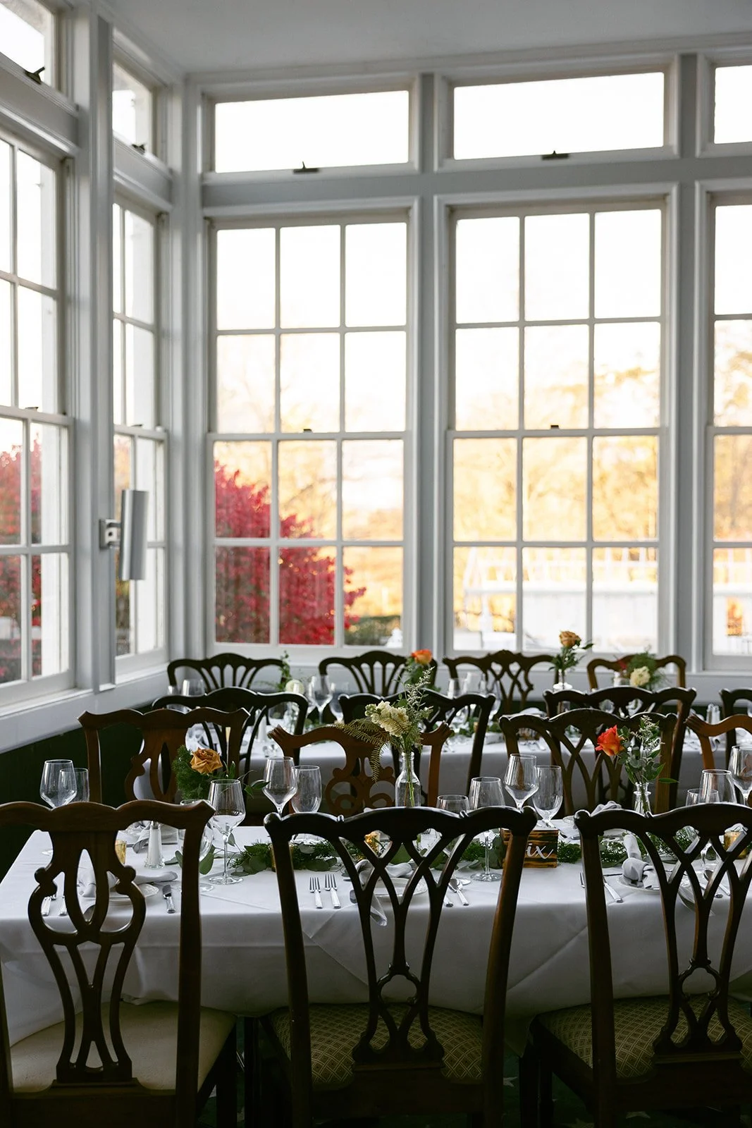Elegant dining room with wooden chairs, white tablecloths, and tables set with glassware, cutlery, and floral centerpieces, beside large sunlit windows.