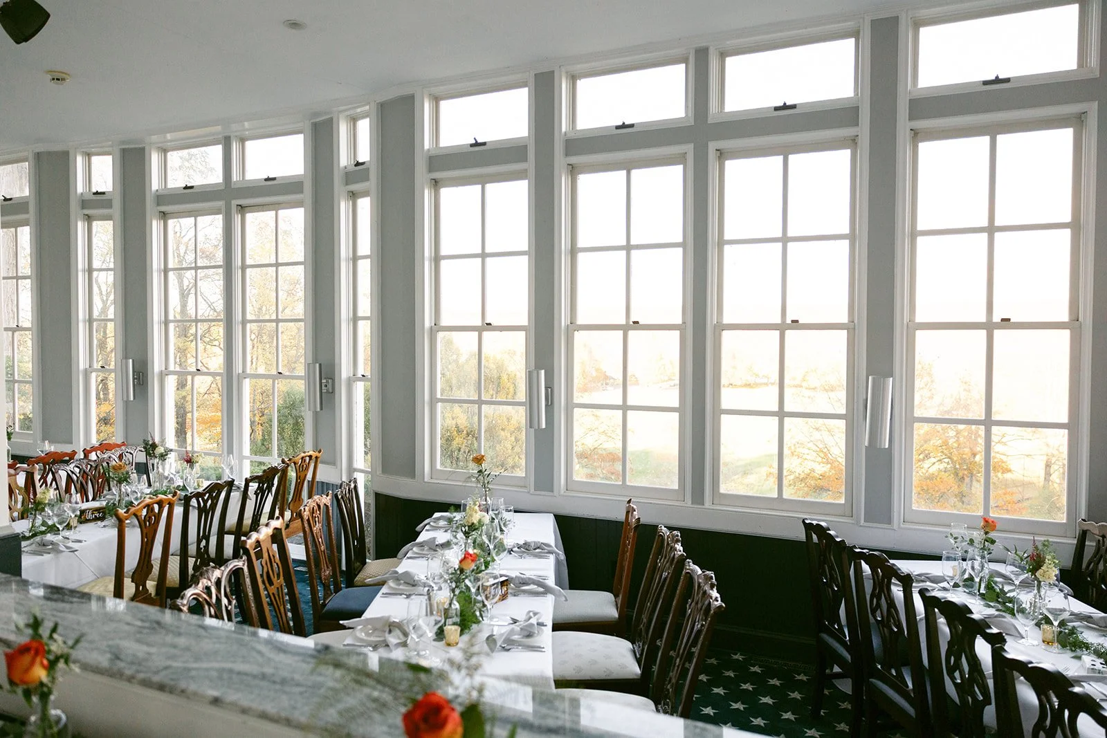 Elegant dining room with wooden chairs, white tablecloths, and tables set with glassware, cutlery, and floral centerpieces, beside large sunlit windows.