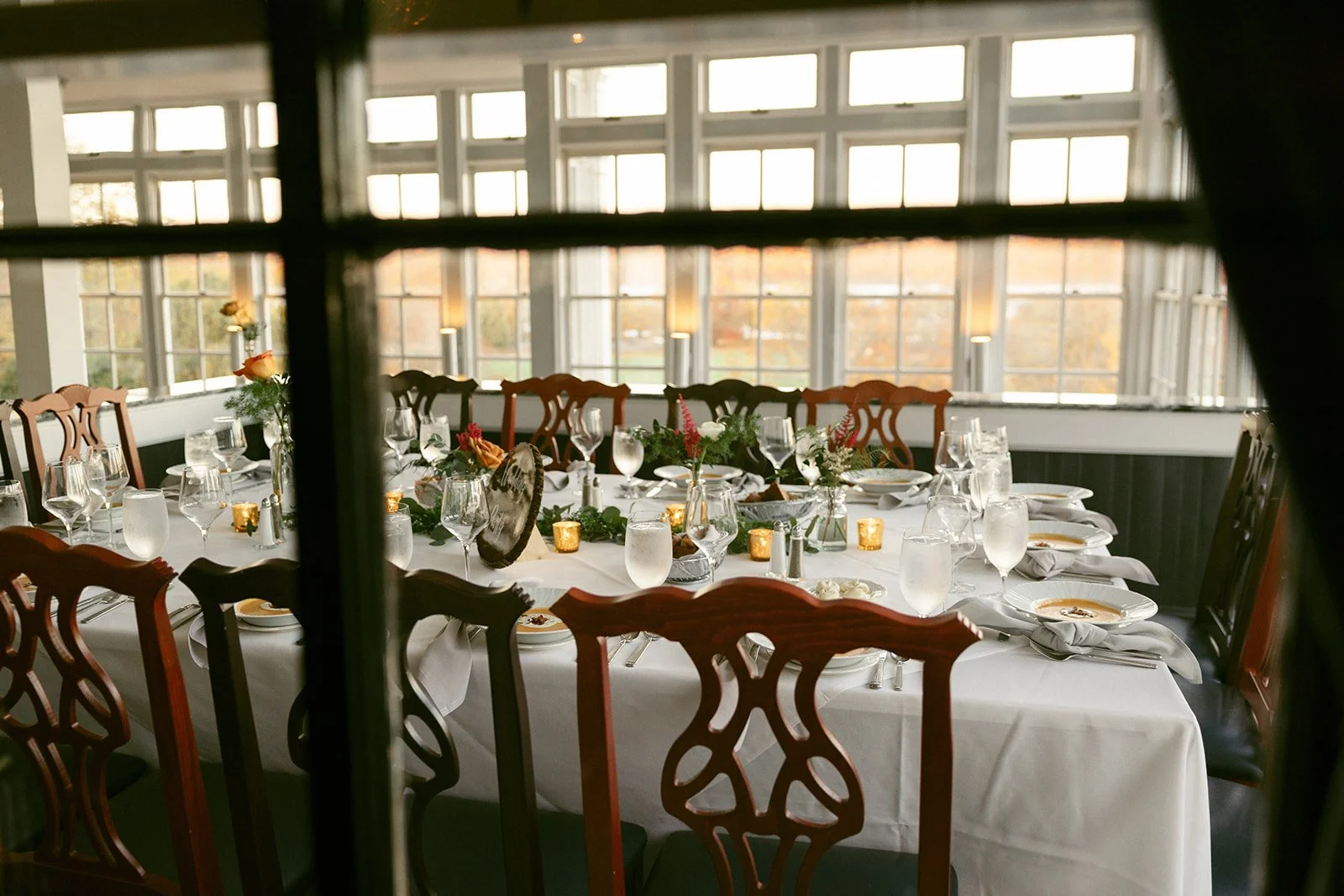 Elegant dining room with wooden chairs, white tablecloths, and tables set with glassware, cutlery, and floral centerpieces, beside large sunlit windows.