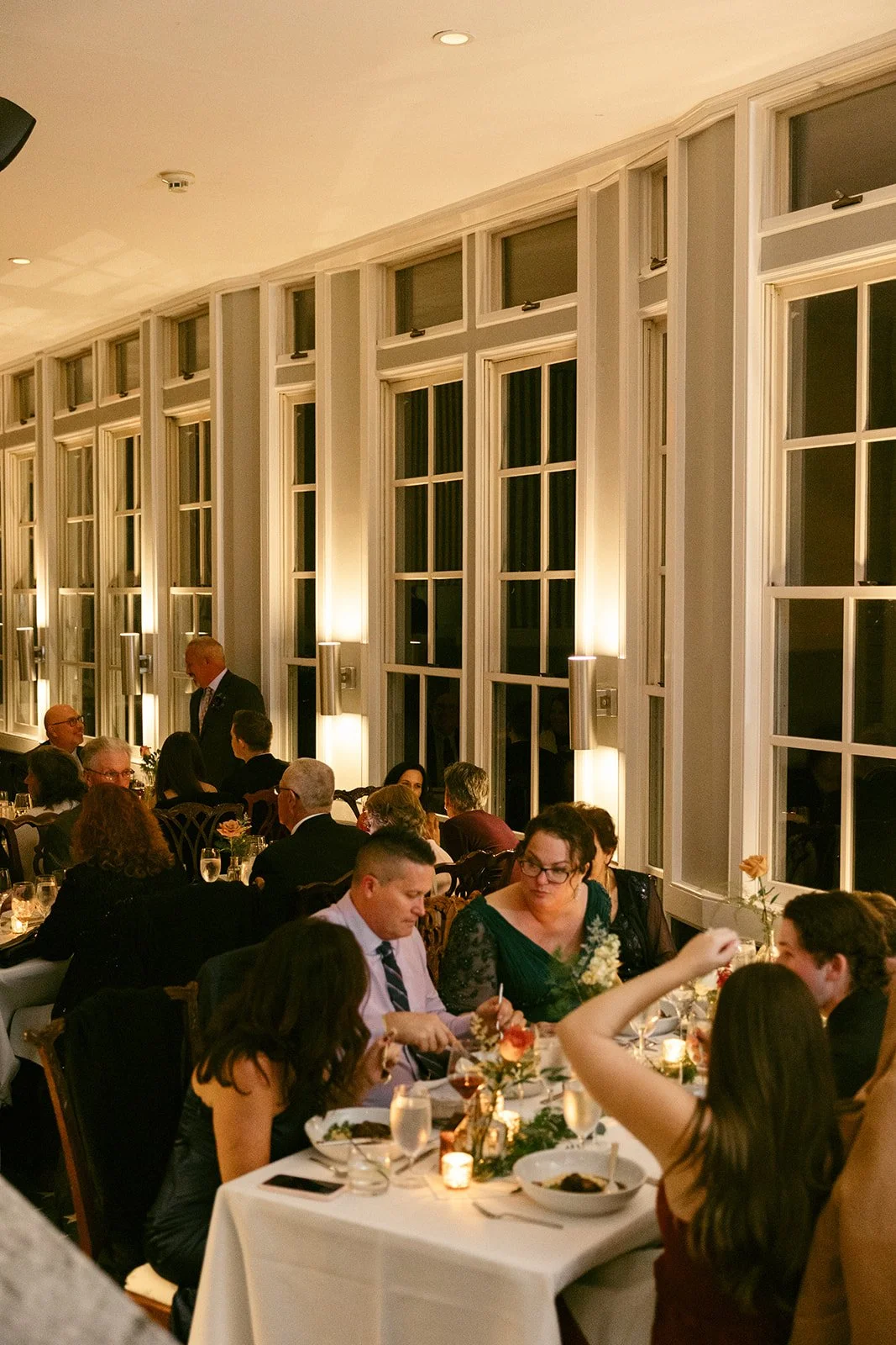 Elegant dining room with wooden chairs, white tablecloths, and tables set with glassware, cutlery, and floral centerpieces, beside large sunlit windows.