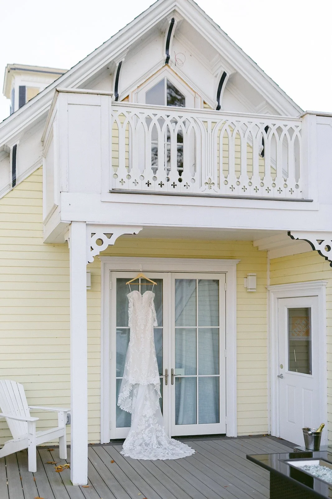 A white lace wedding dress hangs on a hanger from a balcony railing above glass double doors on the porch of a light yellow house.
