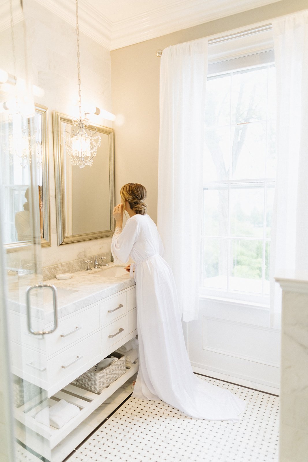 A woman in a white robe stands at a bathroom sink, looking into a mirror, with natural light coming through a large window.