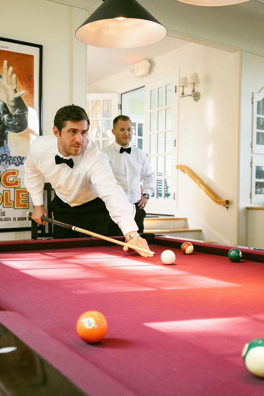 Two men in white shirts and bow ties stand by a red pool table; one is preparing to take a shot while the other looks on.