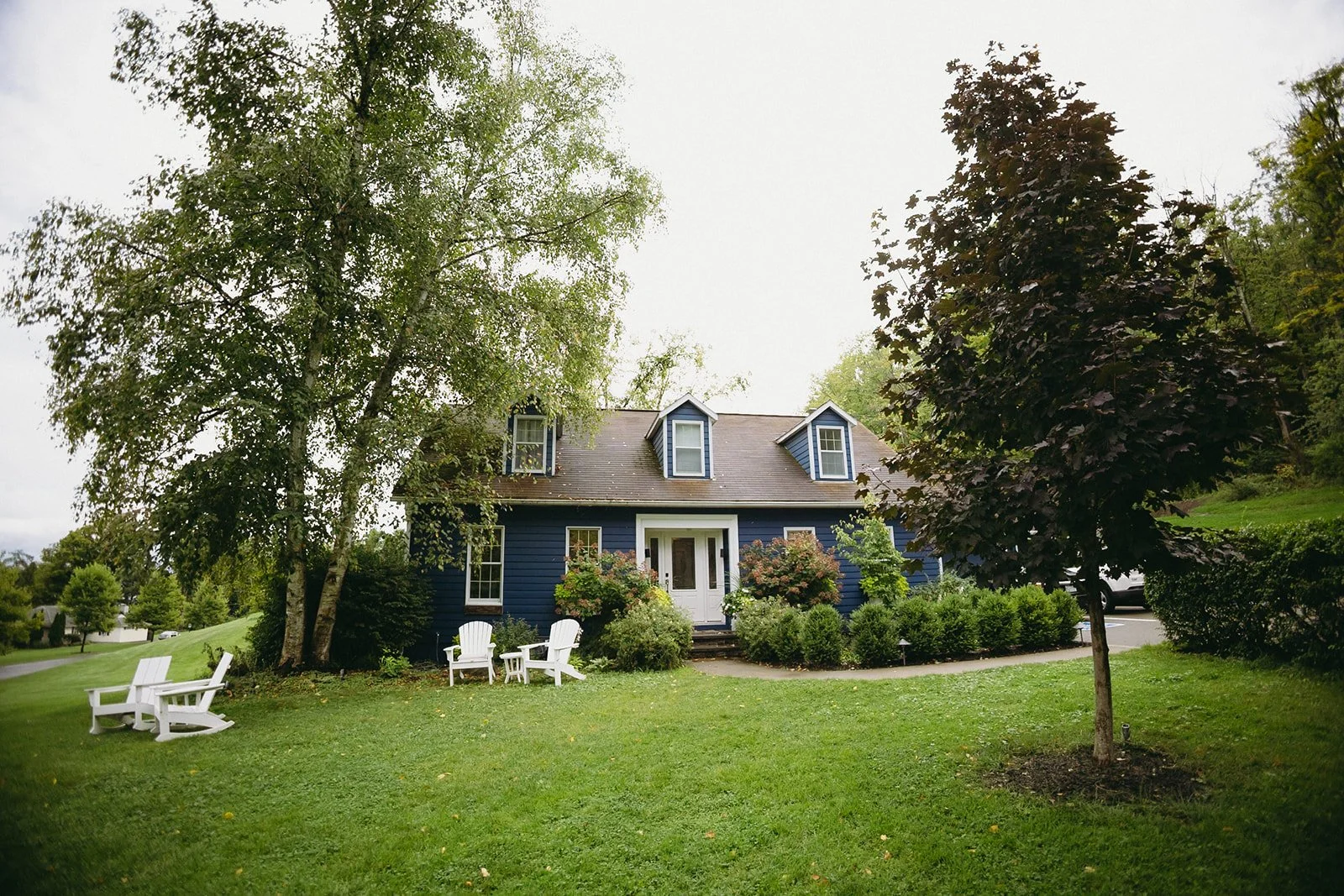A blue house with dormer windows is surrounded by trees, green lawn, and white outdoor chairs arranged near the front.