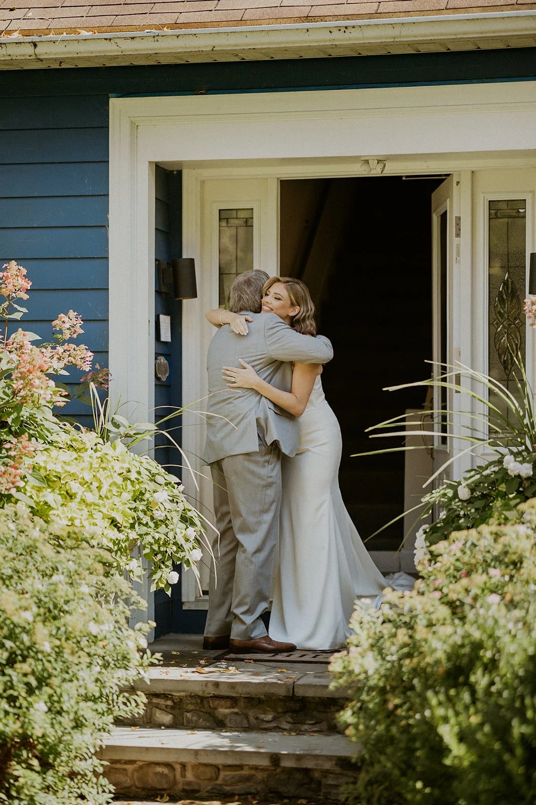 A woman in a white dress hugs an older man in a light suit at the entrance of a house surrounded by greenery.