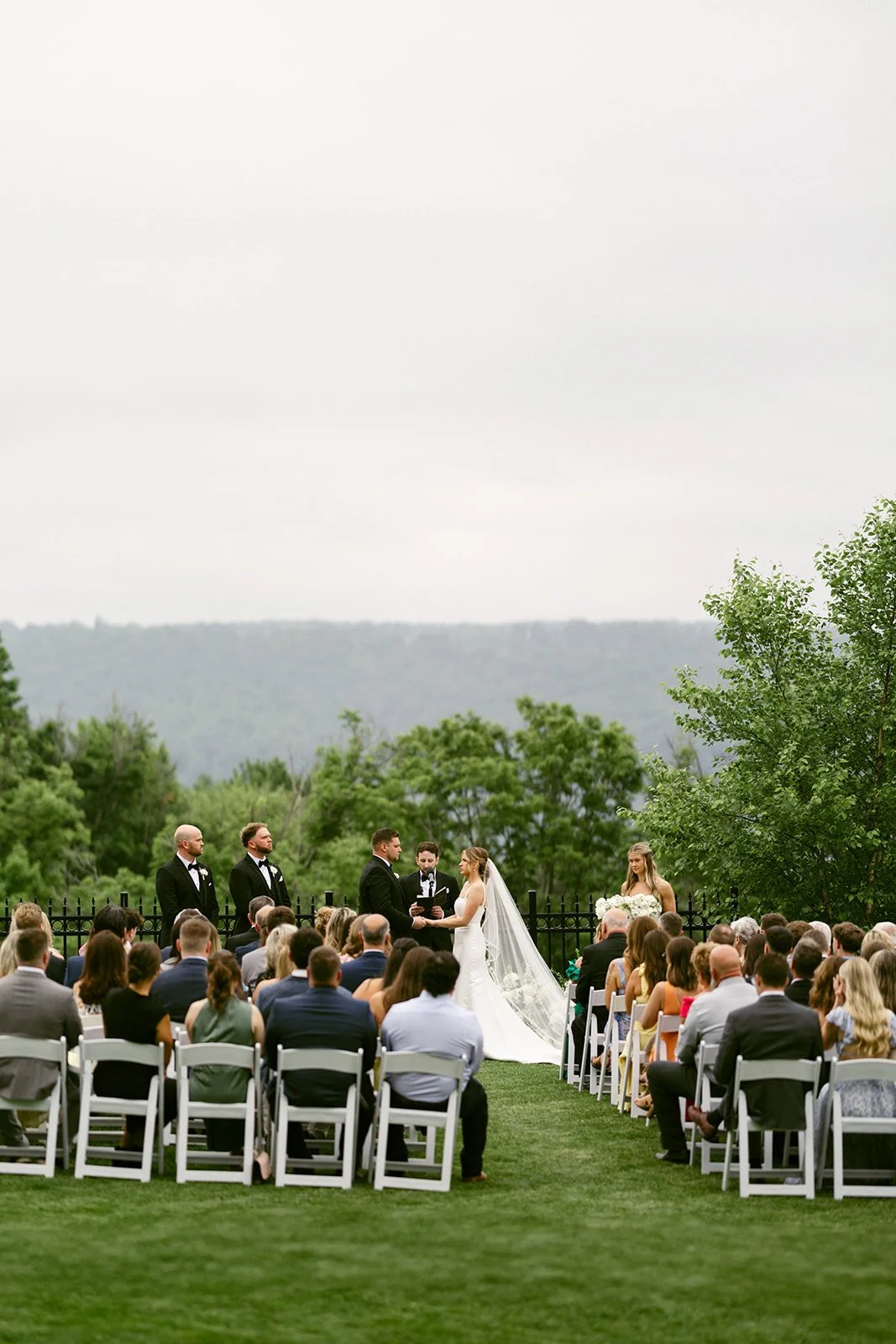An outdoor wedding ceremony with guests seated on white chairs, a couple at the altar, officiant, and bridal party, with trees and hills in the background at for a Inn at Taughannock Falls Wedding