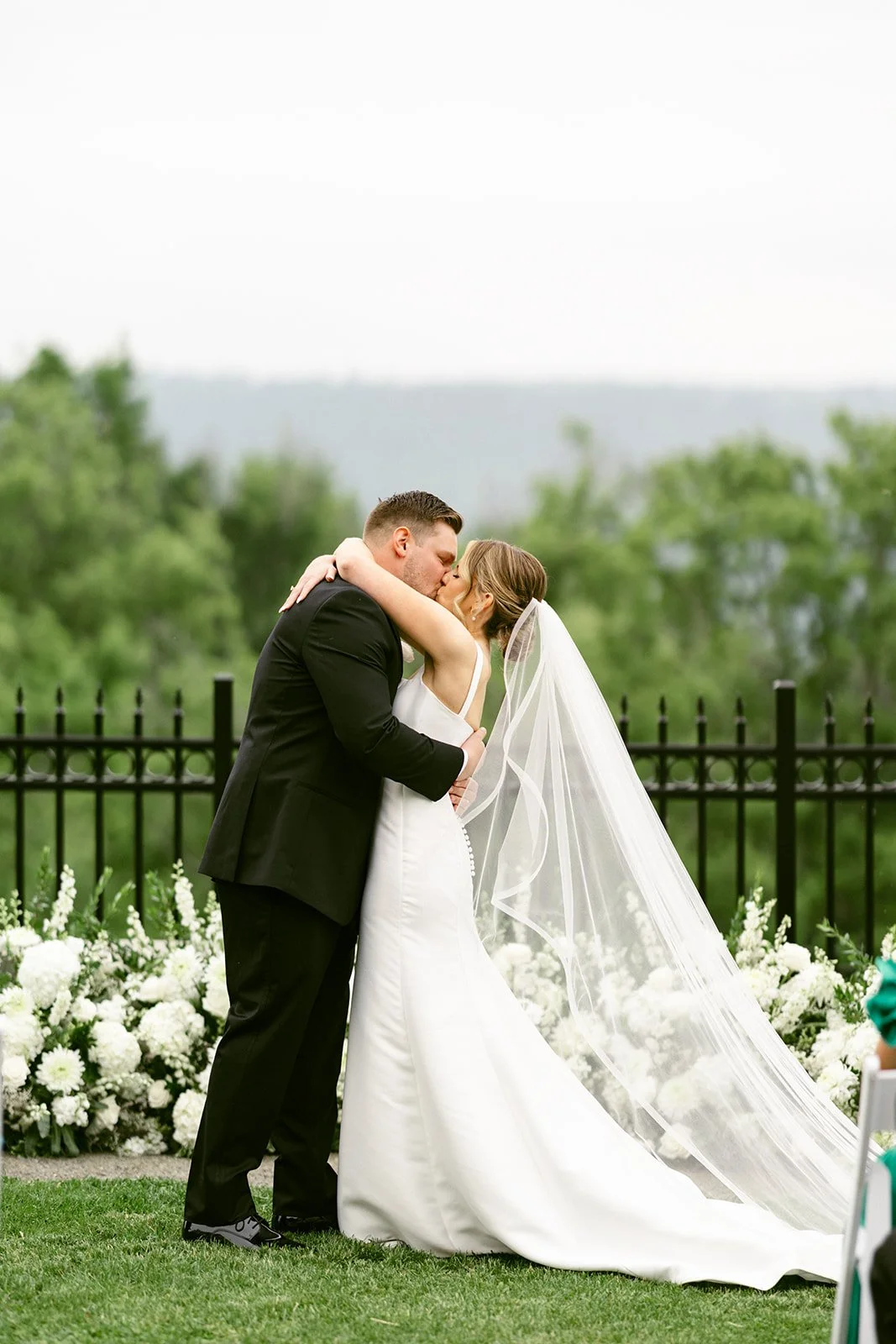 An outdoor wedding ceremony with guests seated on white chairs, a couple at the altar, officiant, and bridal party, with trees and hills in the background at for a Inn at Taughannock Falls Wedding