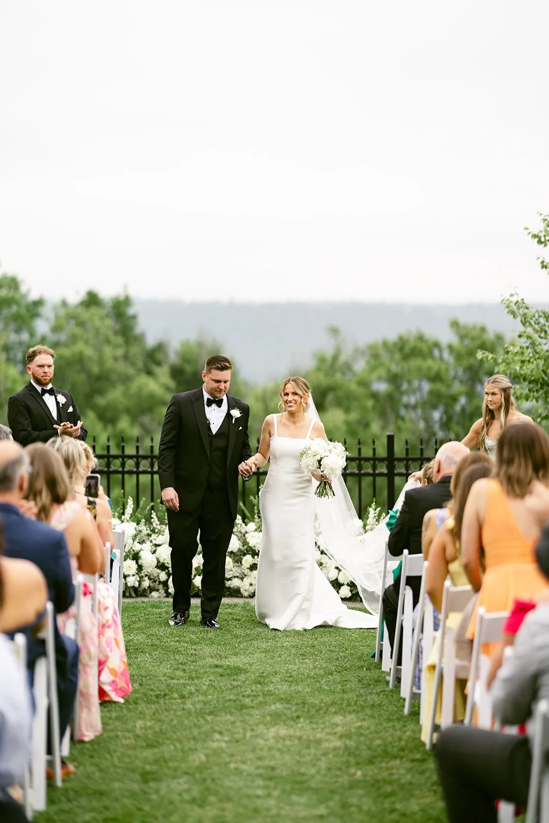 An outdoor wedding ceremony with guests seated on white chairs, a couple at the altar, officiant, and bridal party, with trees and hills in the background at for a Inn at Taughannock Falls Wedding