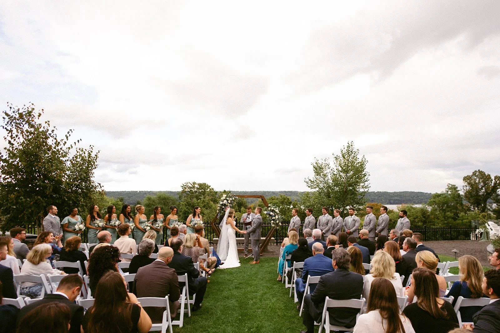An outdoor wedding ceremony with guests seated on white chairs, a couple at the altar, officiant, and bridal party, with trees and hills in the background at for a Inn at Taughannock Falls Wedding