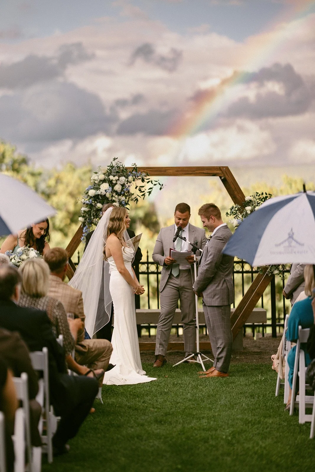 An outdoor wedding ceremony with guests seated on white chairs, a couple at the altar, officiant, and bridal party, with trees and hills in the background at for a Inn at Taughannock Falls Wedding