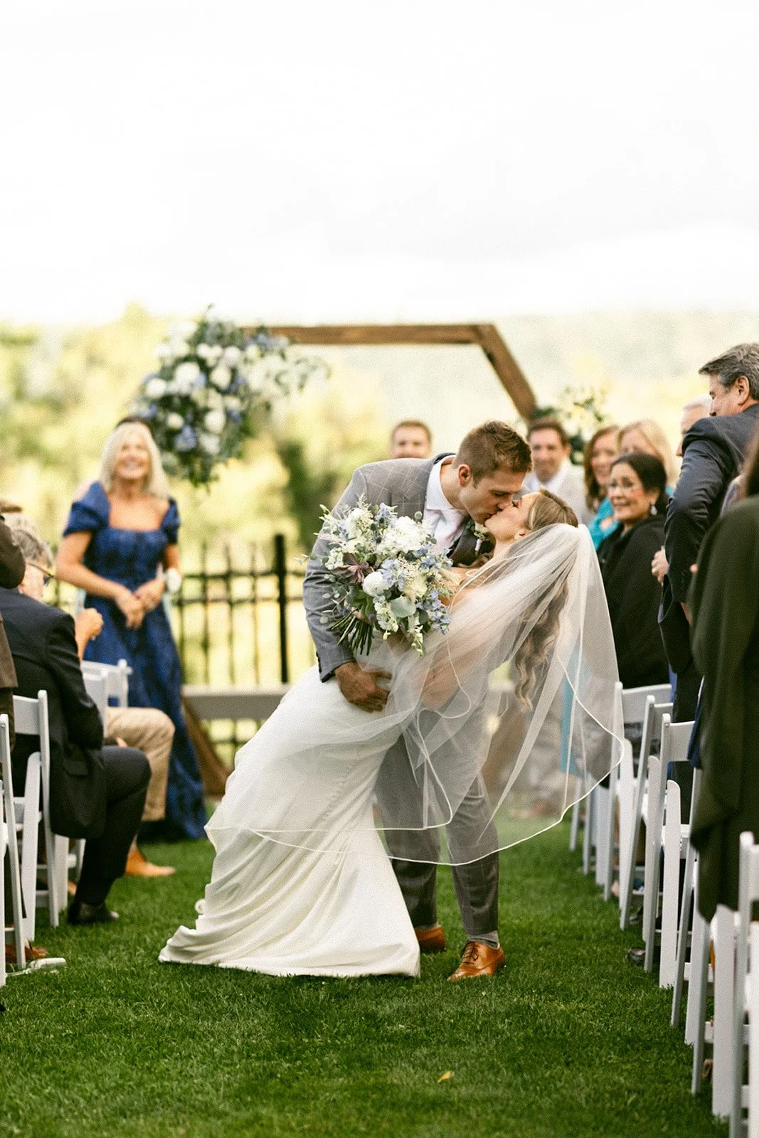 An outdoor wedding ceremony with guests seated on white chairs, a couple at the altar, officiant, and bridal party, with trees and hills in the background at for a Inn at Taughannock Falls Wedding