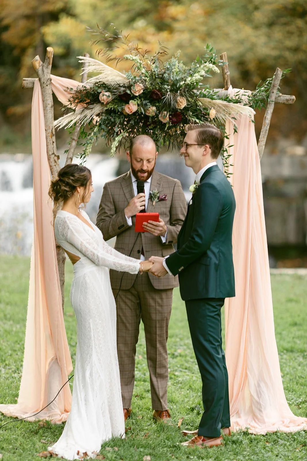 A couple holds hands and faces each other during an outdoor wedding ceremony, with an officiant standing between them under a decorated wooden arch.