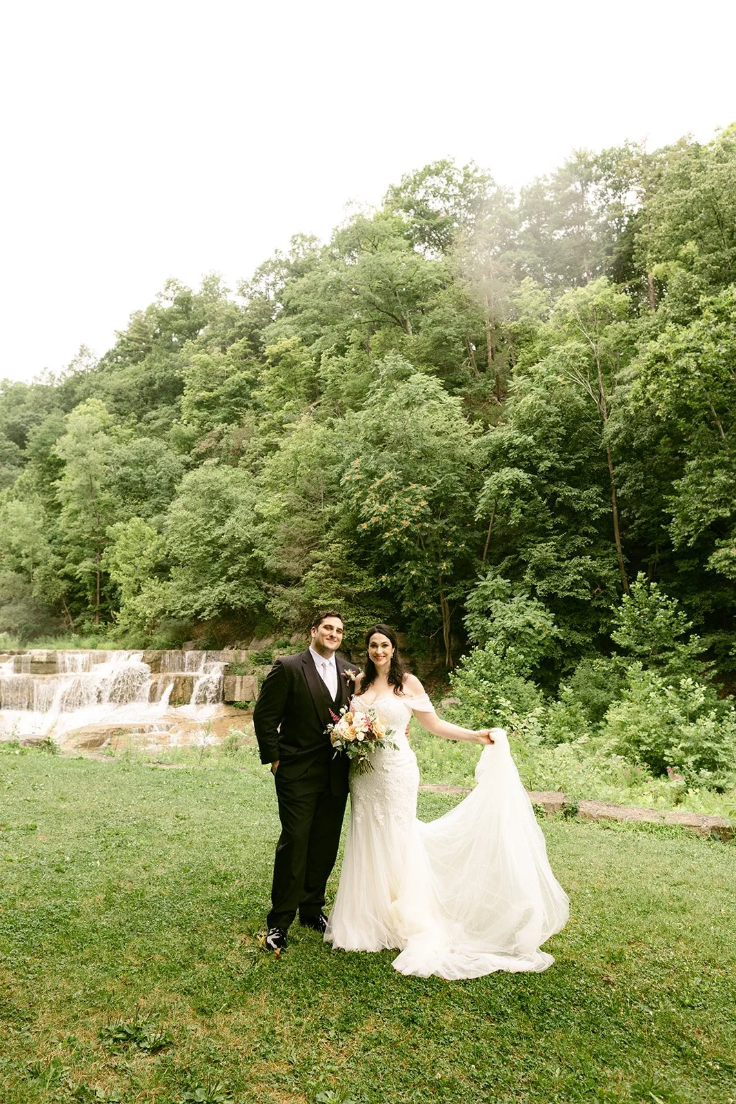 A bride and groom stand together outdoors on grass, with a forest and small waterfall in the background.