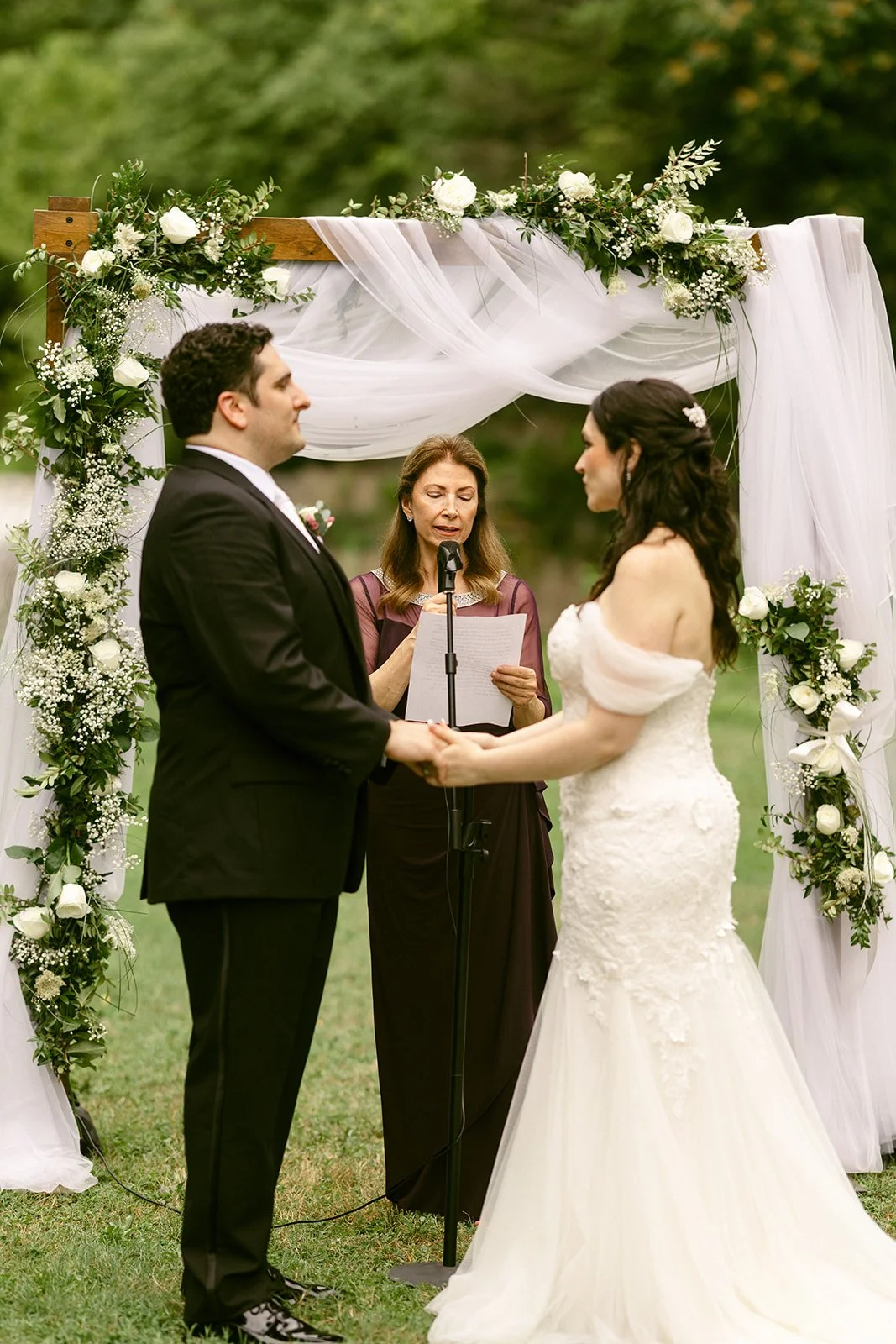 A bride and groom stand together outdoors on grass, with a forest and small waterfall in the background.