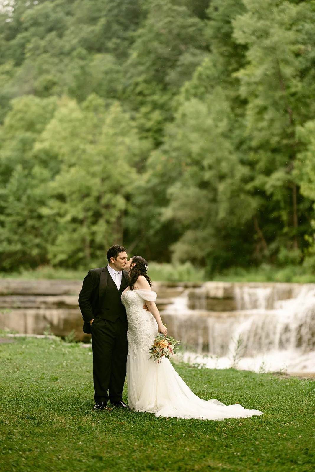 A bride and groom stand together outdoors on grass, with a forest and small waterfall in the background.