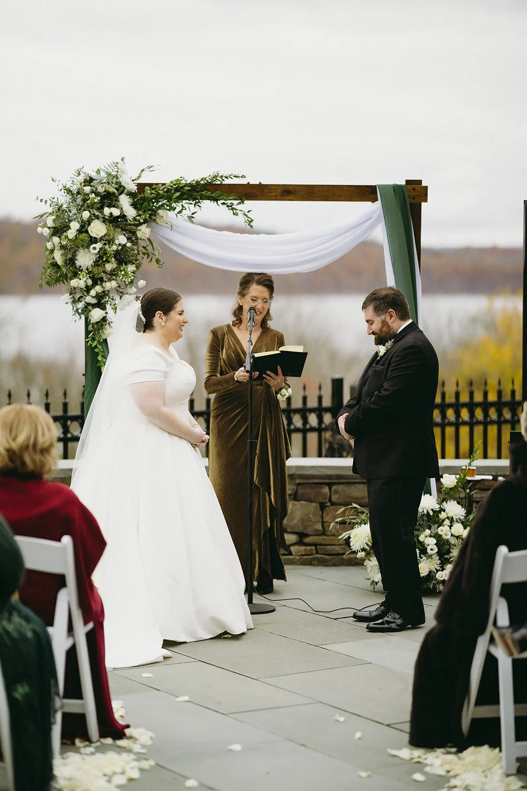 A bride and groom stand before an officiant under a floral arch during an outdoor wedding ceremony, with seated guests in the foreground for an Inn at Taughannock Falls Wedding