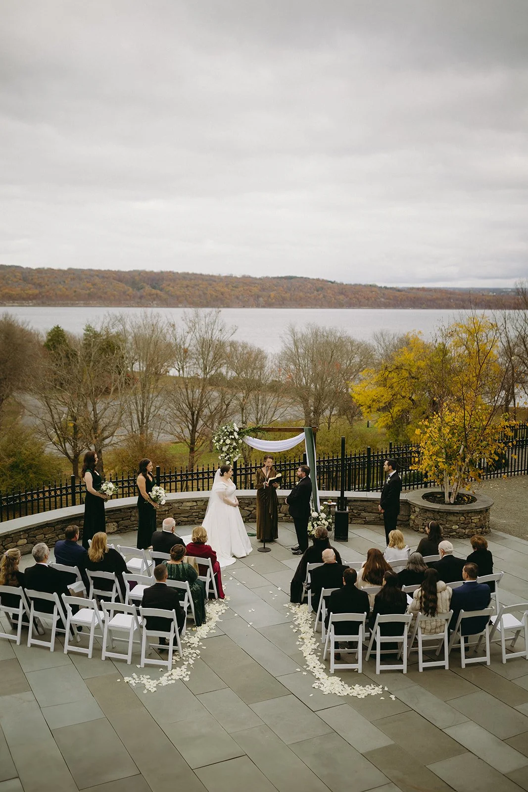 A bride and groom stand before an officiant under a floral arch during an outdoor wedding ceremony, with seated guests in the foreground for an Inn at Taughannock Falls Wedding